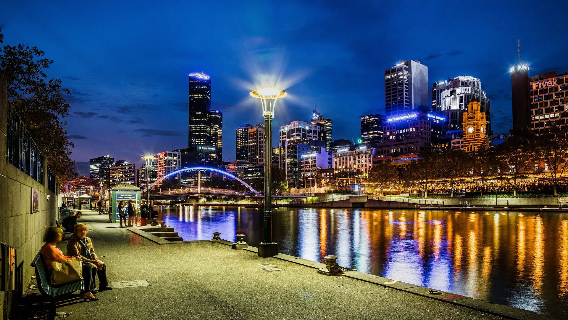 La promenade de Southbank au bord de la Yarra, de nuit, avec un lampadaire, une passerelle éclairée de bleu, des tours illuminées et le clocher de Flinders Street Station se reflétant dans le fleuve.