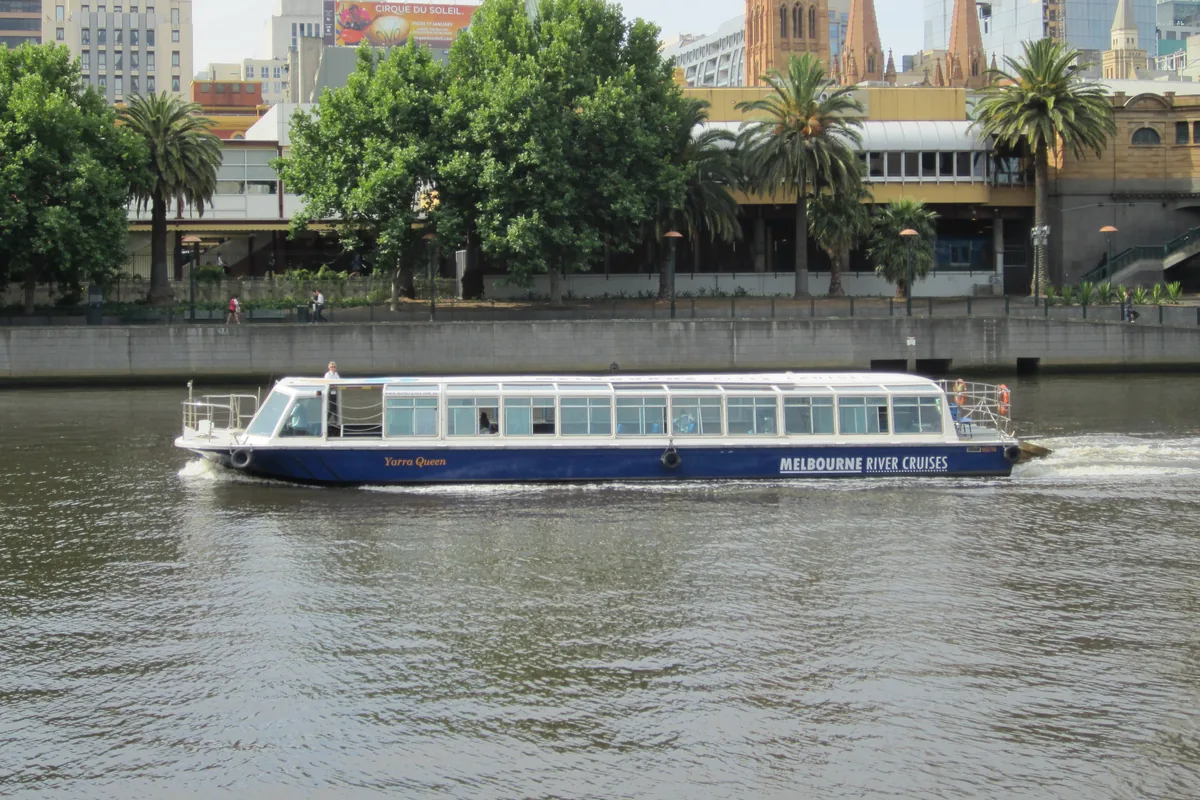Un bateau de croisière fluviale blanc et bleu sur la Yarra, auprès des arbres et des toits des bâtiments riverains de la ville.